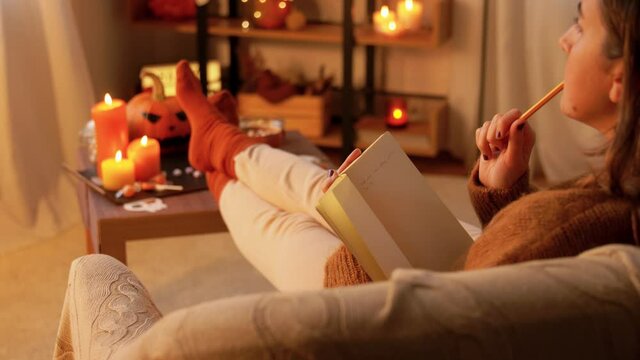 people, holidays and leisure concept - young woman with pencil writing to diary and resting her feet on table at cozy home
