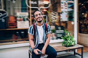 Half length portrait of smiling dark skinned hipster guy sitting on bench with smartphone for...