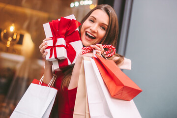 Young excited beautiful woman hold shopping bags, gift boxes and cane candy in hands and laugh outdoors. Cheerful attractive girl shopper with present box, paper bags. Christmas shopping, winter sale