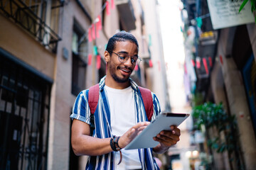 Positive handsome dark skinned hipster guy in eyewear reading notification from navigation app walking on street, smiling african american male traveler searching information about destinations