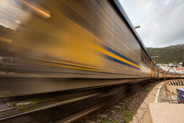 Fototapeta premium Wide angle view of a moving train at the Kalkbay Rail Road crossing in Cape Town in the Western Cape of South Africa