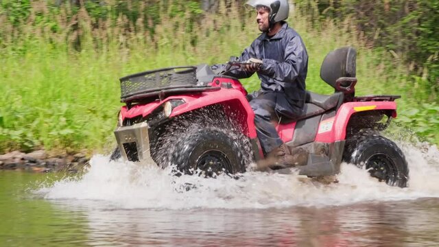 Tracking of bearded man in helmet driving red ATV through river in forest on sunny day