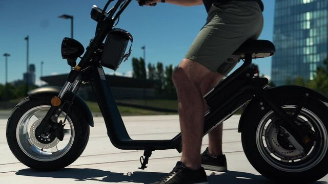 Young man in shorts walking to electric motorcycle and drives away at beautiful location