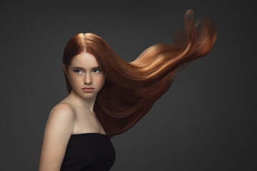 Youth. Beautiful model with long smooth, flying red hair isolated on dark grey studio background. Young caucasian girl with well-kept skin and hair blowing on air. Concept of salon care, beauty