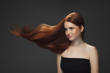 Sea wind. Beautiful model with long smooth, flying red hair isolated on dark grey studio background. Young girl with well-kept skin and hair blowing on air. Concept of salon care, beauty, fashion.