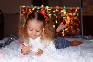a little girl with two ponytails in a white sweater lies on a white rug and draws against the background of Christmas lights