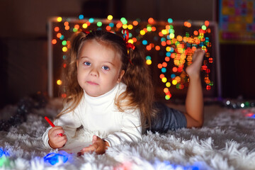 Fototapeta na wymiar a little girl with two ponytails in a white sweater lies on a white rug and draws against the background of Christmas lights