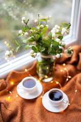 A white snowberry in a bouquet stands with two cups of coffee on the windowsill near the window, beautiful lights, bokeh in the window.