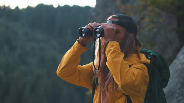 So amazing forest! Bearded tourist man with long hair in yellow jacket wearing backpack looking through binoculars while standing at the top of the hill and inspired by beautiful view in mountain
