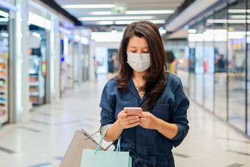 Asian woman using smartphone while wearing mask and walking at shopping mall.