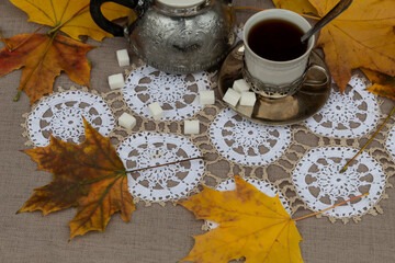 View from above. On a white napkin, among the autumn leaves, there is a cup of tea in a cup holder and a metal teapot. There are sugar cubes nearby.