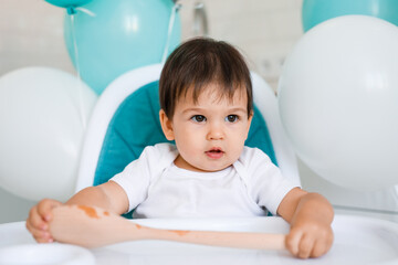 Fototapeta na wymiar Little baby boy sitting in blue high chair at home on white kitchen and playing with wooden big spoon on background with balloons.