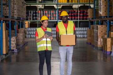 Woman worker using a tablet to check stock online and a man holding a parcel in the automotive parts warehouse distribution. Both engineers people wear safety helmet. In background shelves with goods