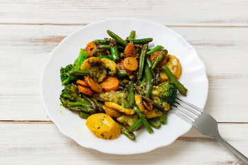Assorted fried vegetables on a plate on a white wooden background. Mix of carrots, green beans, cauliflower.