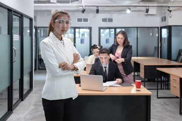 Fototapeta premium Young asian businesswoman confident with crossed arms wearing face shield and coworkers seriously working on desk at modern office