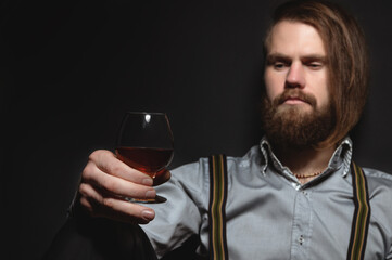 Stylish bearded man in a shirt with suspenders sniffs a glass of whiskey. studio portrait
