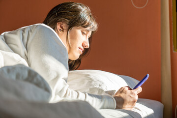 Young woman looking at cellphone in bed after waking up late on weekend