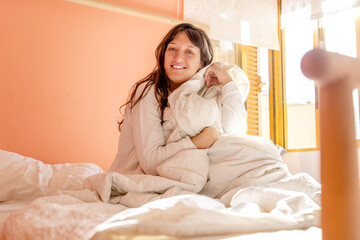 Young woman sitting on the bed dressed in white getting up late with natural light through the window, woman looks and smiles at camera