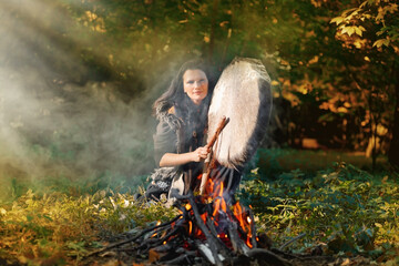 Shaman woman drumming next to the fire in the forest in the setting sun