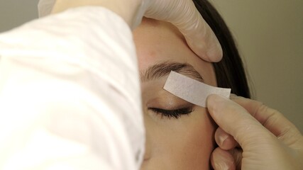 Beautician removes warm wax excess hair from the eyebrow with a special strip. Wax correction of...
