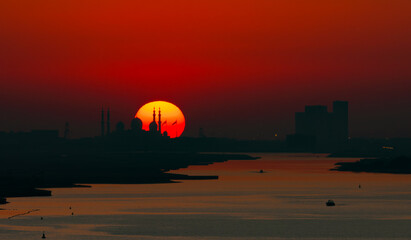 As the sun sets and illuminates the sky in red and in the foreground you see silhouette of the grand mosque with the sun in yellow orange on the horizon and a deep red purple sky