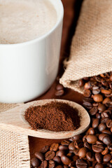 cup of coffee, wooden spoon with ground coffee, raffia cloth sack, brown background