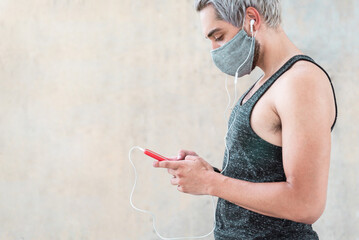 Sport young man listening music playlist on mobile phone while wearing earphones and protective face mask - Coronavirus lifestyle