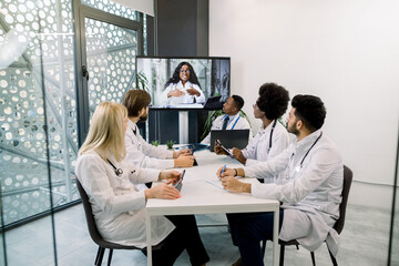 Team of five multiethnical doctors having video conference meeting in hospital with their young African woman doctor. Health care team on webinar meeting online. Telemedicine concept.