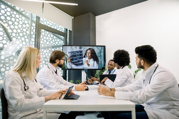 Group of multiethnic doctors scientists sitting at the table and listening to their young African female colleague on computer monitor, discussing computed tomography results during online conference