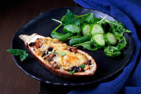 Stuffed eggplant half with melted cheese, mushroom and vegetables roasted and served on the black plate with fresh salad. Low key photo