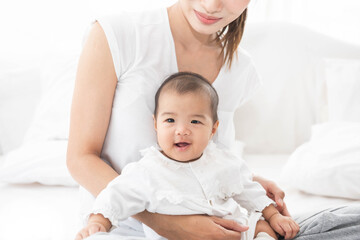 Asian mother and baby laugh together at home. Asia mom holdind infant and sitting on white bed in living room at house  having fun together.