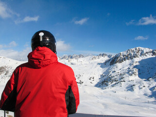 A man in a red jacket and a black ski helmet looks at the white snow peaks and the bright blue sky. Travel content