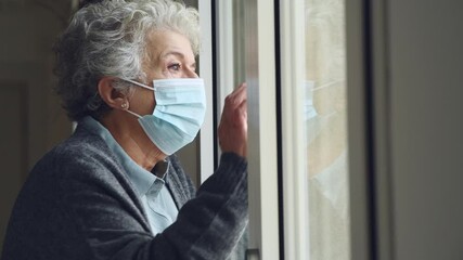 Senior woman with face mask stay at home during Covid-19 pandemic. Depressed old aged woman looking outside of a window with surgical mask during quarantine and lockdown measures for Covid19. - Powered by Adobe