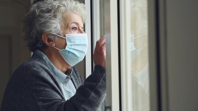 Senior woman with face mask stay at home during Covid-19 pandemic. Depressed old aged woman looking outside of a window with surgical mask during quarantine and lockdown measures for Covid19.