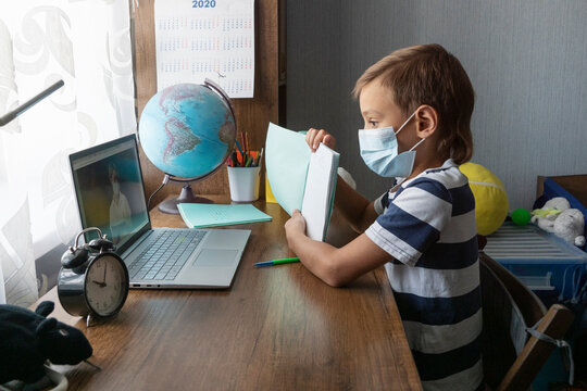 schoolboy in medical face mask shows homework in his notebook to teacher on laptop  screen during online lesson.  Distance education during lockdown.