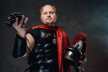 Status and handsome roman general in black armour and red cape with wreath on his bald head poses with grape in smokey background.