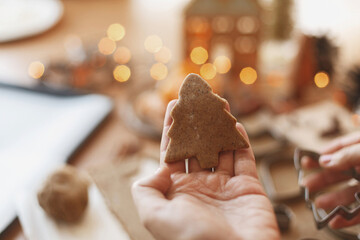 Person holding tree gingerbread cookie on background of festive lights on rustic table. Christmas