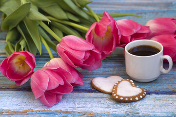 On a blue wooden background: pink tulips, a cup of coffee, heart-shaped cookies. Greeting card for March 8.