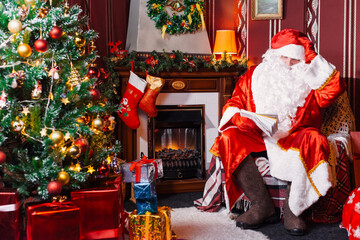 Portrait of Santa Claus - with a white beard, Santa's hat, with gifts near the Christmas tree and fireplace on an armchair on Christmas Eve and New Year.