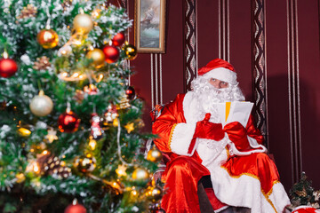 Portrait of Santa Claus - with a white beard, Santa's hat, with gifts near the Christmas tree and fireplace on an armchair on Christmas Eve and New Year.