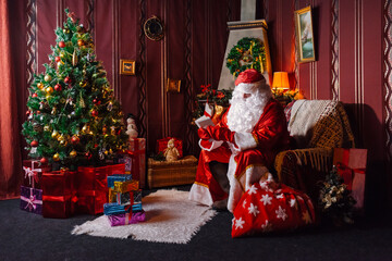 Portrait of Santa Claus - with a white beard, Santa's hat, with gifts near the Christmas tree and fireplace on an armchair on Christmas Eve and New Year.