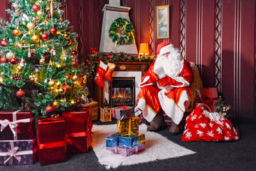 Portrait of Santa Claus - with a white beard, Santa's hat, with gifts near the Christmas tree and fireplace on an armchair on Christmas Eve and New Year.