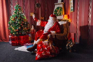 Portrait of Santa Claus - with a white beard, Santa's hat, with gifts near the Christmas tree and fireplace on an armchair on Christmas Eve and New Year.