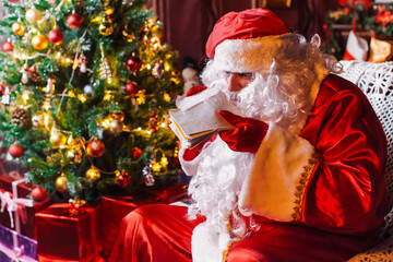 Portrait of Santa Claus - with a white beard, Santa's hat, with gifts near the Christmas tree and fireplace on an armchair on Christmas Eve and New Year.