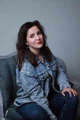 Obraz na płótnie Canvas attractive brunette in jeans and denim jacket sitting on grey chair. studio shoot. 