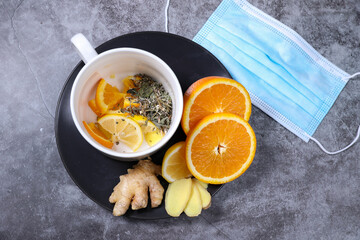 White cup of tea with lemon, ginger, orange., jam and medical mask on the dark grey background. Healthcare. Vitamin cocktail.