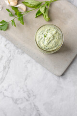 Homemade Green Goddess Dressing in a Jar on a Cement Cutting Board on White Marble Countertop; Parsley and Garlic in Background