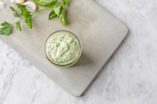 Homemade Green Goddess Dressing in a Jar on a Cement Cutting Board on White Marble Countertop; Parsley and Garlic in Background