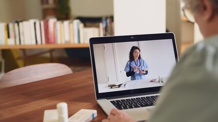 Back view of senior woman making video call with her doctor while staying at home. Close up of patient sitting at table video conferencing with general practitioner on laptop computer. Sick old lady. - Powered by Adobe