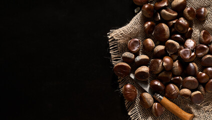 Fototapeta premium Raw chestnuts on burlap on a black background with copy space, preparing for baking. Top view. Chestnut for christmas or holidays, seasonal dish